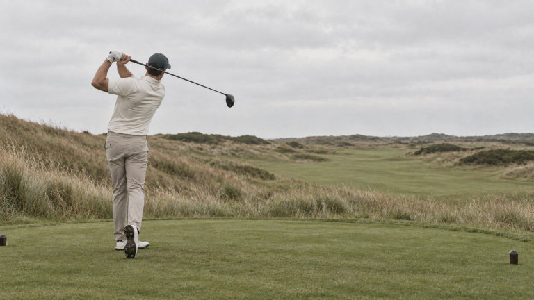 A golfer drives from a tee box on a windswept links course
